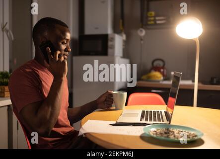 Homme noir avec une tasse de boisson chaude parlant au téléphone tout en travaillant à distance depuis chez vous Banque D'Images