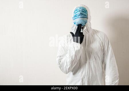 un homme dans un costume de protection accroché à des masques médicaux posant sur un fond de mur montrant divers gestes avec son doigts Banque D'Images