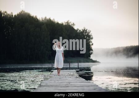 Jeune femme en robe blanche sur un lac le matin froid avec une brume sur l'eau. Banque D'Images