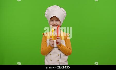 Drôle de caucasien preschooler enfant fille habillé cuisinier chef boulanger dans un tablier et chapeau avec fourchette en plastique et couteau se tromper autour, faisant des visages sur fond de clé chroma. École de cuisine, éducation des enfants Banque D'Images