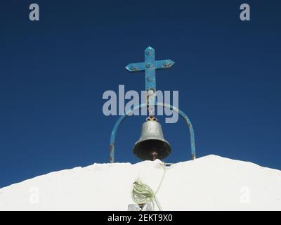 Croix et cloche en métal bleu rouille sur le toit de Une minuscule église orthodoxe grecque traditionnelle blanche avec du bleu clair fond ciel Banque D'Images
