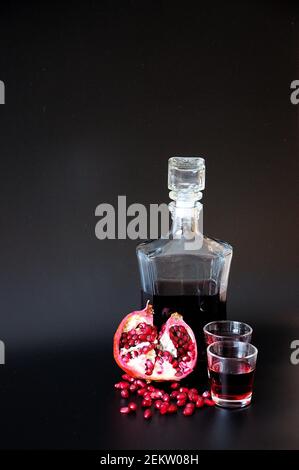 Une bouteille de verre et deux verres de liqueur de grenat se tiennent sur un fond noir à côté des fruits brisés et des graines. Positionnement vertical. Banque D'Images