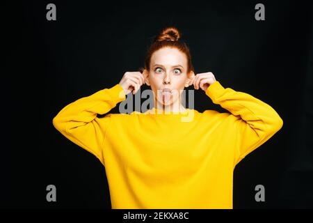 Folle joyeuse jeune femme en chandail jaune touchant les oreilles avec mains et la langue Banque D'Images