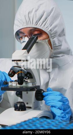 Une chercheuse porte une combinaison d'epi et regarde des échantillons au microscope dans un laboratoire équipé. Médecin travaillant avec diverses bactéries, tissus, analyses de sang, recherche pharmaceutique pour les antibiotiques Banque D'Images