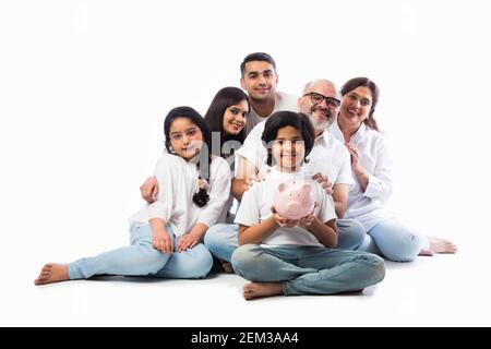 Famille indienne multigénérationnelle de six personnes tenant une tirelire tout en portant des chiffons blancs et se tenant contre un mur blanc Banque D'Images