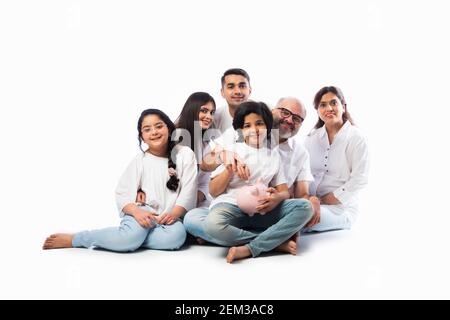 Famille indienne multigénérationnelle de six personnes tenant une tirelire tout en portant des chiffons blancs et se tenant contre un mur blanc Banque D'Images