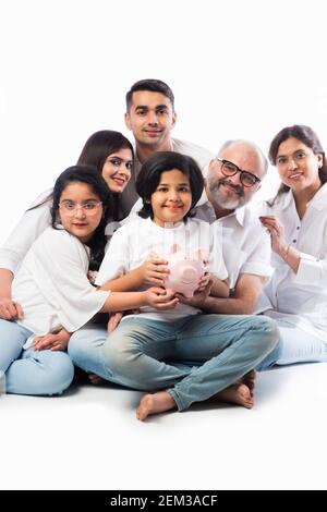 Famille indienne multigénérationnelle de six personnes tenant une tirelire tout en portant des chiffons blancs et se tenant contre un mur blanc Banque D'Images