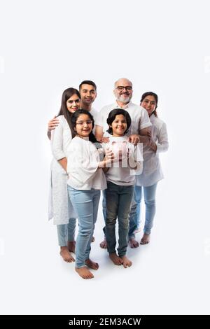 Famille indienne multigénérationnelle de six personnes tenant une tirelire tout en portant des chiffons blancs et se tenant contre un mur blanc Banque D'Images