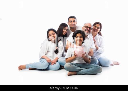 Famille indienne multigénérationnelle de six personnes tenant une tirelire tout en portant des chiffons blancs et se tenant contre un mur blanc Banque D'Images