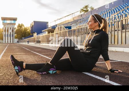 femme se reposant après avoir courez au stade. Fitness femme portrait style de vie en noir tracksuit extérieur. Coucher de soleil sur fond, magnifique perspec Banque D'Images