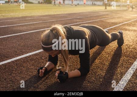 Jeune femme athlétique faisant de l'exercice de planche sur la piste de course à pied. Coucher de soleil sur fond, belle perspective. Banque D'Images