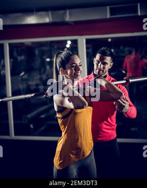 Jeune souriant sain sportif forte forme active fille tenant barbell sans poids sur l'épaule à l'avant tout en étant utile personnel formateur debout Banque D'Images