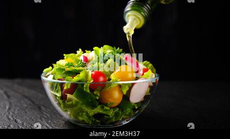Huile d'olive versé de la bouteille sur la salade de légumes sur le noir arrière-plan Banque D'Images