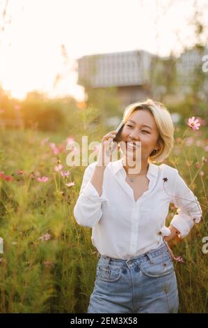 Jeune belle fille asiatique debout dans un champ vert avec des fleurs, souriant heureusement, parlant sur son téléphone, chaud sentiment insouciant au coucher du soleil. Banque D'Images