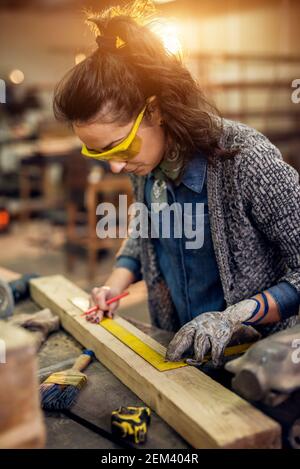 Vue rapprochée de la femme charpentier professionnelle sérieuse et concentrée tenir la règle et le crayon tout en faisant des marques sur le bois à la table de la Banque D'Images