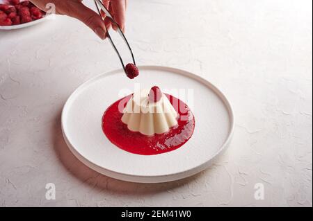 La main de femme met des pinces framboises à la Panna cotta avec du sirop, clé haute, gros plan Banque D'Images