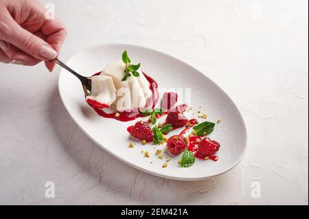 La main d'une femme tient une cuillère avec un morceau de panna cotta avec sirop, framboises et menthe dans une assiette ovale, le style de la nourriture, clé haute, gros plan Banque D'Images