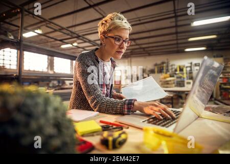 Gros plan de cheveux courts focalisés attrayant industriel d'âge moyen femme ingénieur avec lunettes tenant des plans tout en travaillant un ordinateur portable au travail Banque D'Images