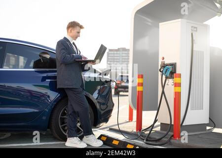 Jeune homme beau travaillant sur un ordinateur portable, attendant pendant que sa voiture électrique de luxe est charching, payer pour la charge, se tenant près de la gare de ville avec l'électricité Banque D'Images