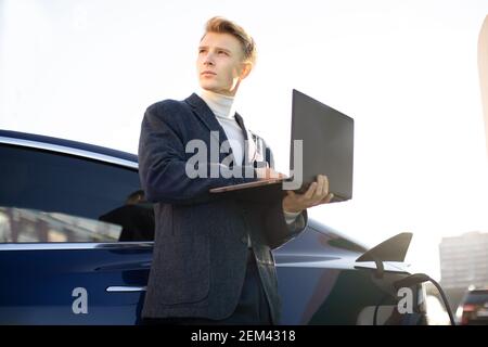 Jeune homme d'affaires européen charmant à la station de charge, debout près d'une voiture électrique avec un ordinateur portable et regardant loin, tout en ravitaillant sa voiture. VÉ Banque D'Images