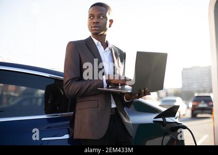 Homme d'affaires noir à la station de charge, debout près d'une voiture électrique avec un ordinateur portable et regardant loin, tout en ravitaillant sa voiture. Station EV et Banque D'Images