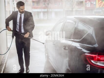 Vue portrait d'un jeune homme plein de confiance et plein de style homme en costume lavant sa voiture noire avec de l'eau canon sur l'auto-analyseur manuel Banque D'Images