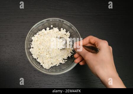 La main d'une femme avec une cuillère prend un grain de fromage cottage d'un bol en verre sur une table noire, vue de dessus Banque D'Images