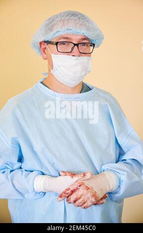 Portrait d'un médecin masculin dans des lunettes regardant l'appareil photo. Homme chirurgien portant des gants stériles, un uniforme chirurgical bleu, un capuchon médical, un masque de protection. Concept de la médecine, des travailleurs de la santé et de la chirurgie. Banque D'Images