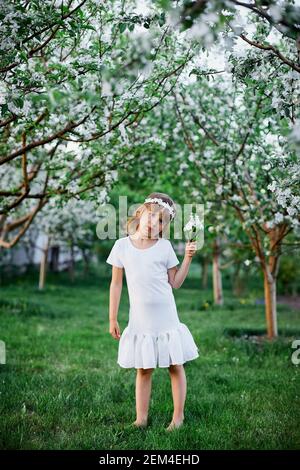 Adorable petite fille de 5-6 ans tenant une fleur debout dans le jardin de printemps de fleur, portant une robe blanche et une couronne florale à l'extérieur, la saison du printemps est à venir. Banque D'Images