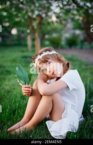 Adorable petite fille de 5-6 ans tenant la fleur est assis dans le jardin de printemps de fleur, portant la robe blanche et la couronne florale dehors, la saison de printemps est à venir Banque D'Images