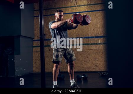 Gros plan au-dessus de la vue forte motivé et concentré musculaire homme de bodybuilder barbu de cheveux courts tenant deux grands kettlebells rouges devant pendant le train Banque D'Images