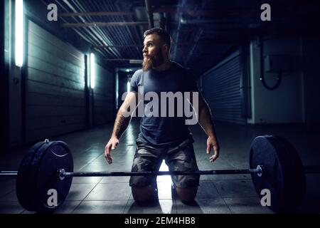 Vue rapprochée de la forme physique active et résistante bodybuilder barbu homme s'agenouillant sur le sol avant de soulever la barbell lourde le métro Banque D'Images