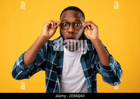 Noir émerveillé Homme regardant l'appareil photo à travers des lunettes, fond jaune Banque D'Images