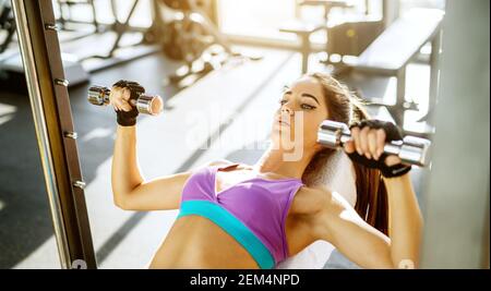 Gros plan horizontal de concentré motivé mince actif sain jeune femme faisant de l'exercice de poitrine avec de petites haltères sur le banc dans le mod ensoleillé Banque D'Images