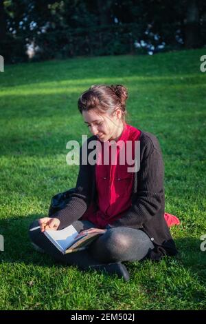 Fille assise sur l'herbe dans le parc public, lit un livre et fume une cigarette. Banque D'Images