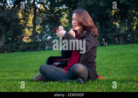 Fille assise sur l'herbe dans le parc public, met sur le maquillage et met sur le rouge à lèvres. Banque D'Images