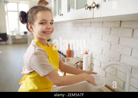 Petite fille souriante dans un tablier debout laver la vaisselle cuisine à la maison Banque D'Images