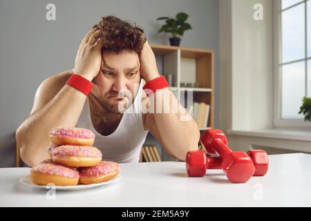 Triste homme regardant les haltères et les beignets choisir entre sain et un style de vie malsain Banque D'Images