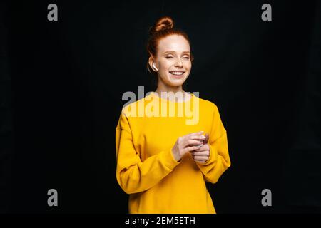 Une jeune femme en train de rire, portant des écouteurs sans fil, écoutant des chansons populaires arrière-plan noir isolé Banque D'Images