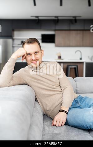 Homme confiant et mince avec des cheveux bruns clairs, des yeux gris et de courtes soies propres, assis, tête reposant sur une main et regardant l'appareil photo, flirting, amical, posant, portant le chandail beige et le Jean Banque D'Images