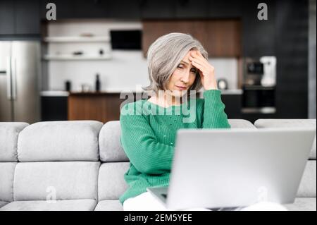 Contrarié femme senior regarde l'écran de l'ordinateur portable et se sent frustré, gagner à la loterie, a un problème avec le travail. Une dame à la retraite mature reçoit une mauvaise nouvelle ou ne peut pas se permettre des factures Banque D'Images