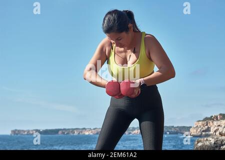 Femme latine d'âge moyen, avec dessus jaune, léopard noir, avec haltères, gymnastique, réchauffé pour l'entraînement, brûler des calories, garder la forme, en plein air par mer, Banque D'Images