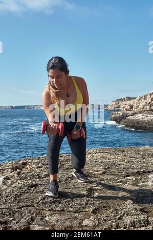 Femme latine d'âge moyen, avec dessus jaune, léopard noir, avec haltères, gymnastique, réchauffé pour l'entraînement, brûler des calories, garder la forme, en plein air par mer, Banque D'Images