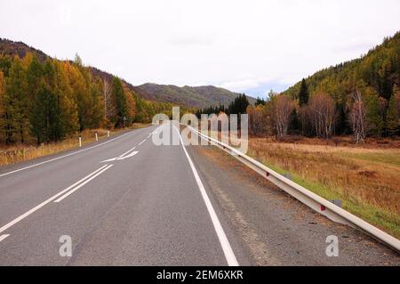 Une route asphaltée à deux voies avec une clôture passe au loin à travers une pittoresque vallée d'automne. Région de Chuisky, Altaï, Sibérie, Russie. Banque D'Images