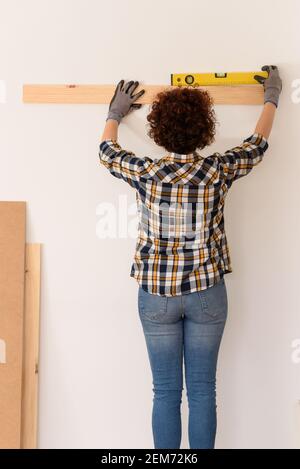 une femme méconnaissable utilise un niveau pour placer avec précision une planche en bois sur un mur blanc à l'intérieur d'un plat baigné de lumière naturelle. Banque D'Images