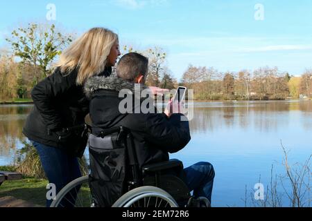 Concept d'une personne handicapée. Un homme en fauteuil roulant avec une femme debout à côté de lui. Couple utilisant la technologie tout en regardant un smartphone. Banque D'Images