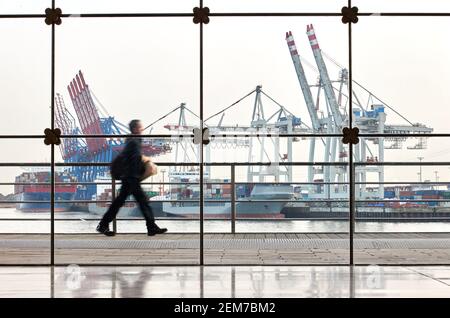 Hambourg, Allemagne. 22 février 2021. Un homme marche derrière une façade en verre à Holzhafen d'Altona. Les navires à conteneurs et les grues du terminal à conteneurs de Tollerort sont visibles en arrière-plan. Le 25.02.2021, la conférence de presse annuelle en ligne du port de Hambourg sur le développement du port de Hambourg et le transport maritime de marchandises en 2020 a lieu. (Effet de suppression dû à une longue exposition) crédit : Georg Wendt/dpa/Alay Live News Banque D'Images