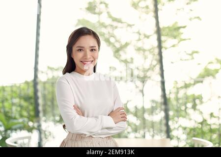 Jeune heureuse belle femme asiatique avec des tences, bras debout croisés, souriant à l'appareil photo dans un bureau moderne avec grande fenêtre en verre et vert flou Banque D'Images