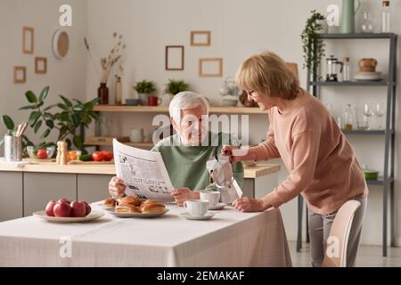 Femme âgée versant du thé chaud pour son mari pendant qu'il assis à la table et lisant le journal pendant le petit-déjeuner la cuisine Banque D'Images