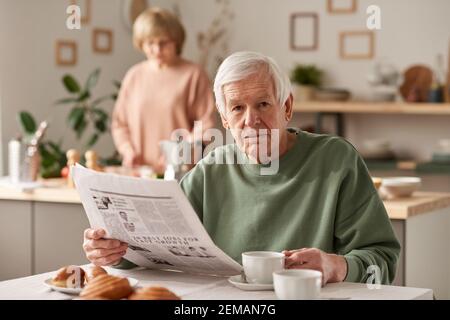 Portrait d'un homme âgé regardant l'appareil photo tout en lisant un journal pendant le petit déjeuner dans la cuisine Banque D'Images